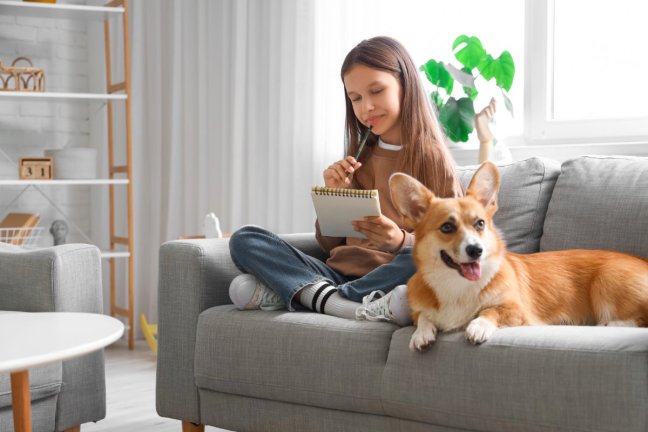 girl writing in a notebook, sitting on couch with her corgi